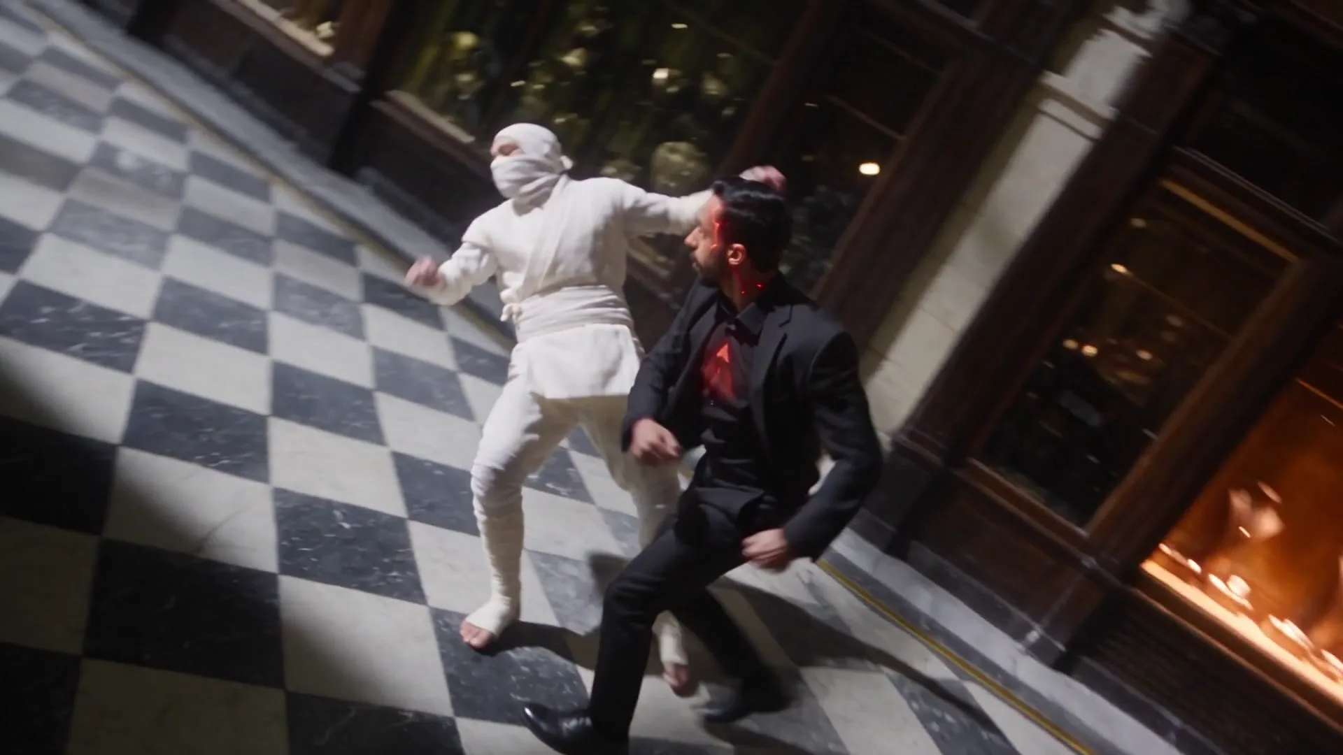 Photogramme du film : dans un passage couvert parisien de nuit, au sol de marbre en damier, un adversaire en blanc frappe un homme en costume noir dont les veines du cou et l'emblème au sternum rougeoient comme de la braise.